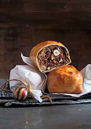 Ein angeschnittenes Früchtebrot mit dunklem Kern und dünnem, hellem Mantel liegt auf einem Früchtebrot im Ganzen und einem in Pergamentpapier eingewickelten Früchtebrot. Im hellen Kern des Früchtebrotes sind Haselnüsse und Walnüsse in der Fruchtmasse erkennbar.