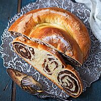 Eine halbe Gubana mit glänzender, goldbraun ausgebackener, feiner Kruste liegt auf einer dunklen Platte mit weißem Rankenmuster. Die Krume der Gubana zeigt noch die Windungen der Teigrolle mit der dunklen Frucht-Nuss-Füllung, die zur Schnecke gelegt gebacken wurde.