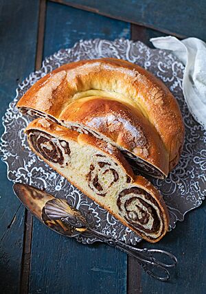Eine halbe Gubana mit glänzender, goldbraun ausgebackener, feiner Kruste liegt auf einer dunklen Platte mit weißem Rankenmuster. Die Krume der Gubana zeigt noch die Windungen der Teigrolle mit der dunklen Frucht-Nuss-Füllung, die zur Schnecke gelegt gebacken wurde.