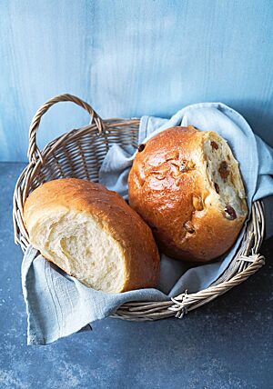 In einem Holzkorb, der mit einem hellgrauen Tuch ausgelegt ist, liegen ein Milchbrot und ein Rosinenbrot. Beide haben eine sehr wattig-weiche Krume und eine goldbraun ausgebackene, glänzende, zarte Kruste.