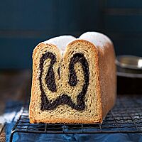Ein angeschnittener Mohnstollen liegt auf einem dunklen Gitterrost. Die hellen und dunklen Schichten verdeutlichen die in der Kastenform gebackene Doppelrolle. Bestreut wurde der Mohnstollen nach dem Backen mit Puderzucker.