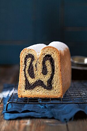 Ein angeschnittener Mohnstollen liegt auf einem dunklen Gitterrost. Die hellen und dunklen Schichten verdeutlichen die in der Kastenform gebackene Doppelrolle. Bestreut wurde der Mohnstollen nach dem Backen mit Puderzucker.