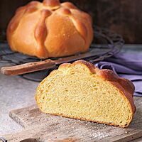 Ein halbes Pan de Muerto mit glatter, glänzender und mit Zucker bestreuter Kruste liegt auf einem alten Holzbrett. Die gelbe Krume ist elastisch und wattig. Im Hintergrund liegt ein weiteres Pan de Muerto.