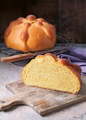 Ein halbes Pan de Muerto mit glatter, glänzender und mit Zucker bestreuter Kruste liegt auf einem alten Holzbrett. Die gelbe Krume ist elastisch und wattig. Im Hintergrund liegt ein weiteres Pan de Muerto.