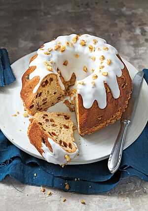 Ein angeschnittener polnischer Babka mit einer dicken Schicht Zuckerguss und gehackten Walnüssen auf der Oberfläche liegt auf einer weißen Platte. Im Anschnitt sind viele Sultaninen in der elastischen, saftigen Krume erkennbar.