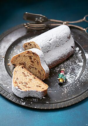 Der mit Puderzucker bestreute Quarkstollen liegt auf einer dunklen, runden Platte. Zwei abgeschnittene Scheiben liegen vor dem Stollen. Sie zeigen die saftige, feinporige Krume.