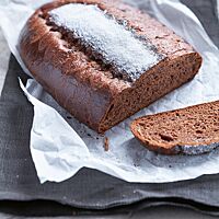 Ein ovaler, flacher Schoko-Brioche-Laib liegt auf einem weißen Pergamentpapier. In der Mitte der Oberfläche ist zwischen einem eingeschnittenen Zackenmuster ein breiter weißer Zuckerstreifen zu erkennen. Die dunkle Krume ist weich, elastisch und kleinporig.