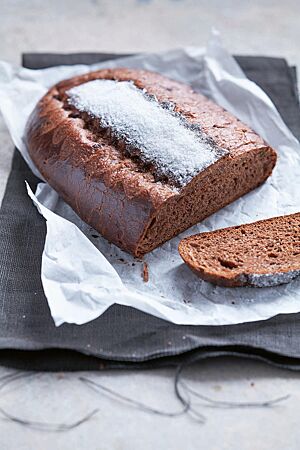 Ein ovaler, flacher Schoko-Brioche-Laib liegt auf einem weißen Pergamentpapier. In der Mitte der Oberfläche ist zwischen einem eingeschnittenen Zackenmuster ein breiter weißer Zuckerstreifen zu erkennen. Die dunkle Krume ist weich, elastisch und kleinporig.