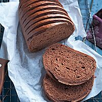 Das angeschnittene Schokobrot liegt auf weißem Pergamentpapier auf einem Gitterrost. Die dunkelbraune Krume ist kleinporig und wattig-weich; die glänzende Kruste ist eng parallel, schräg eingeschnitten.