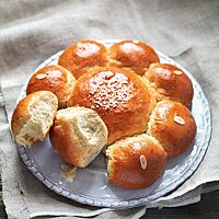 Der blütenförmige Dreikönigskuchen liegt auf einem hellen Teller. Zwei runde Stücke des Dreikönigskuchen liegen abgebrochen an den Kuchen gelehnt und zeigen die faserige, wattig weiche Krume. In der Mitte des Dreikönigskuchen wurde Hagelzucker auf den mit Ei abgestrichenen Teigling gestreut, auf die Oberfläche der kleinen runden Teiglinge ringsum wurde jeweils ein Mandelblättchen gelegt.