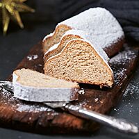 Drei Scheiben des Vollkornmandelstollens liegen auf einem dunklen, rustikalen Holzbrett vor dem mit Feinkristallzucker bestreuten Stollen. Die Krume ist einheitlich hellbraun. Ab und an sind ganze Mandeln in der Krume zu erkennen.