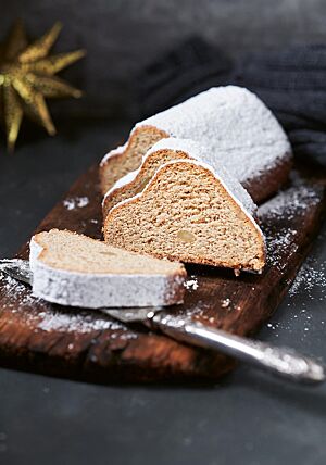 Drei Scheiben des Vollkornmandelstollens liegen auf einem dunklen, rustikalen Holzbrett vor dem mit Feinkristallzucker bestreuten Stollen. Die Krume ist einheitlich hellbraun. Ab und an sind ganze Mandeln in der Krume zu erkennen.