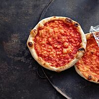 Zwei nur mit Tomatensauce bestrichene Pizzen weisen am Rand der hellen Kruste einige dunkle Stellen auf.