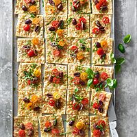 Auf einem Blech liegt die mit Mozzarella, Kirschtomaten, Oliven und Basilikum belegte rechteckige Pizza Romana.