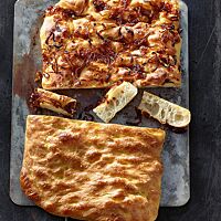 Eine goldbraun ausgebackene Focaccia wurde zur Hälfte mit Zwiebeln belegt (oben) bzw. mit Meersalz bestreut (unten) und liegt halbiert auf einem flachen Blech. Ihre Krume ist luftig-locker, weich und grobporig.