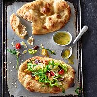 Auf einem Blech liegen eine ovale, goldgelb ausgebackene klassische Pinsa mit Olivenöl und Salz (oben) und eine mit Salat, Tomaten, Oliven und gehobeltem Parmesan belegte Pinsa verde (unten).