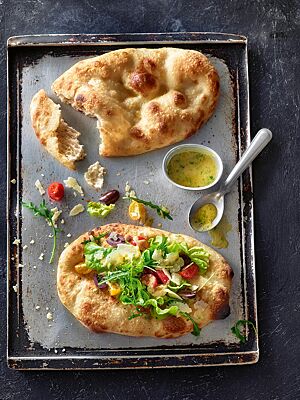 Auf einem Blech liegen eine ovale, goldgelb ausgebackene klassische Pinsa mit Olivenöl und Salz (oben) und eine mit Salat, Tomaten, Oliven und gehobeltem Parmesan belegte Pinsa verde (unten).