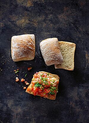 Die hellbraun ausgebackenen Pizzabrötchen haben eine glatte, leicht bemehlte Kruste und eine kleinporige, lockere Krume. Ein halbiertes Pizzabrötchen wurde als Bruschetta mit würzigem Tomatensalat belegt.