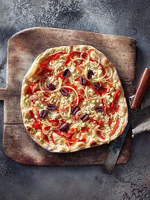 Der knusprig ausgebackene Flammkuchen mit Sauerteig ist mit Crème fraîche, Paprika, Zwiebeln, Oliven und Feta belegt.