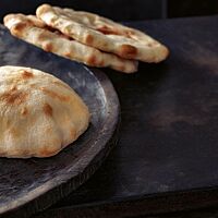 Zwei flache Pitas liegen angelehnt an den Rand eines Teller, auf dem sich eine aufgeblähte Pita befindet. Die kleinen Fladenbrote haben eine helle Krume mit einigen braunen Stellen.