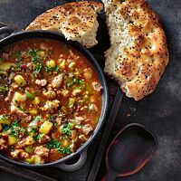 Neben einem Topf mit orientalischer Gulaschsuppe liegt eine große angebrochene Pita mit fluffiger, offenporiger Krume und goldgelb bis goldbraun ausgebackener Kruste.