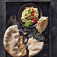 Ein auseinandergebrochenes Aish Baladi mit lockerer, brauner Krume und leicht bemehlter, heller Kruste liegt mit einer Schüssel Baba Ghanoush auf einem dunklen Backblech.