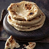 Mehrere kleine runde Kulchas liegen aufeinandergestapelt auf einer runden Holzplatte. Die Fladenbrote sind hell ausgebacken mit einigen dunklen Stellen und haben eine weiche, lockere Krume.