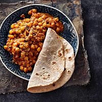 Ein Chapati liegt auf dem Rand einer Schale mit Channah Masala. Das dünne, runde Fladenbrot ist hell ausgebacken mit einigen dunklen Stellen.