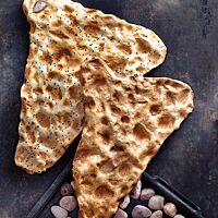 Neben einem Blech mit Kieselsteinen liegt ein Hefeteig-Sangak (oben) und ein Sauerteig-Sangak (unten). Ihre wellige, hell ausgebackene Oberfläche mit vielen goldbraunen Stellen zeugt von den Kieselsteinen, auf denen sie gebacken wurden.