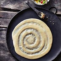 Auf einer Gusseisenplatte liegt ein hellgelb ausgebackener, pfannkuchenähnlicher Anjero-Fladen, auf dessen Oberfläche spiralförmig dicke und dünne Bereiche zu sehen sind. Typischerweise wird er nur von einer Seite gebacken.