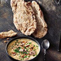 Zwei flache, durchgetrocknete Schüttelbrote lehnen aufrecht an einer Wand neben einer Schüssel Knoblauchrahmsuppe mit Schüttelbrotstückchen.