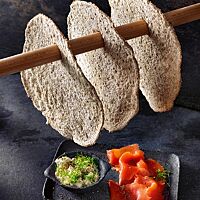 Auf einer kleinen Platte liegen Räucherlachs und mit Kresse bestreuter körniger Frischkäse. Darüber hängt eine Stange, an der drei dünne, runde Knäckebrote aufgehängt sind.