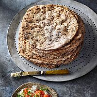 Auf einer runden Platte liegen neben einem Schälchen mit körnigem Frischkäse und Tomaten übereinandergestapelt mehrere runde Flatkaka mit gestippter, leicht bemehlter Oberfläche.