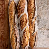 Drei Baguettes mit kräftig ausgebackener, knuspriger Kruste lehnen an einer Holzwand.