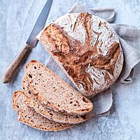 Der angeschnittene Dinkelschmatzer liegt auf einem hellen Untergrund. Im Anschnitt des rustikal aufgerissenen Brotes wird die kleinporige Krume sichtbar. Neben dem Brot liegt ein Brotmesser mit rustikalem Holzgriff.