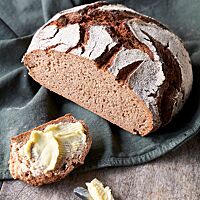 Der Bauernlaib ist ein rundes Brot mit bemehlter, rustikal aufgerissener Kruste und einer lockeren, kleinporigen Krume. Der angeschnittene Laib liegt auf einer grünen Decke. Im Vordergrund liegt eine dick mit Butter bestrichene Brotscheibe.