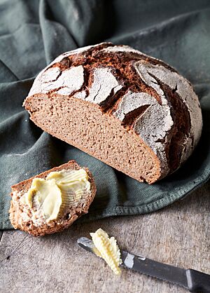 Der Bauernlaib ist ein rundes Brot mit bemehlter, rustikal aufgerissener Kruste und einer lockeren, kleinporigen Krume. Der angeschnittene Laib liegt auf einer grünen Decke. Im Vordergrund liegt eine dick mit Butter bestrichene Brotscheibe.