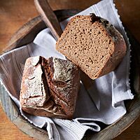 Das Roggen-Kastenbrot liegt halbiert in einer mit einem Tuch ausgelegten Holzschale. Die Kruste des Brotes ist rustikal aufgerissen und die Krume saftig und elastisch.