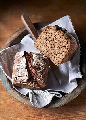 Das Roggen-Kastenbrot liegt halbiert in einer mit einem Tuch ausgelegten Holzschale. Die Kruste des Brotes ist rustikal aufgerissen und die Krume saftig und elastisch.