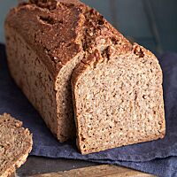 Der angeschnittene, in der Form gebackene Feldhamster liegt auf einem dunkelblauen Tuch. In der an den Laib gelehnten Brotscheibe sind die Roggenkörner in der Krume deutlich sichtbar. Die glänzende Kruste ist rustikal aufgerissen.