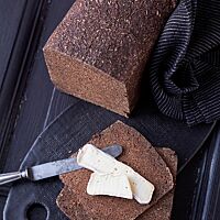 Der angeschnittene Pumpernickel liegt neben dünn geschnittenen Pumpernickelscheiben auf einer dunklen Platte. Im Anschnitt ist die feinporige, kompakte Krume gut sichtbar.