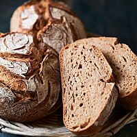 Drei Dreikornbrote mit bemehlter, knuspriger Kruste liegen in einem Holzkorb. Die im Anschnitt sichtbare Krume ist kleinporig und elastisch.