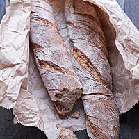 Die beiden Knorrigen mit bemehlter, rustikaler Kruste liegen in einer aufgerissenen, braunen Papiertüte. Das abgebrochene Stück Brot lässt die kleinporige, elastische Krume sichtbar werden