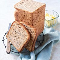 Zwei getoastete Scheiben lehnen an dem angeschnittenen Toastbrot, das hochkant auf einem Gitterrost steht. Im Vordergrund liegt ein rustikales Brotmesser, im Hintergrund steht ein Schälchen mit Butter. Die fluffige, feinporige Krume wird von einer feinen Kruste umgeben.
