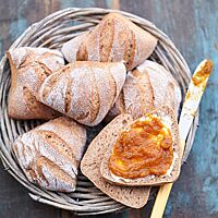 Die eckigen Weizenkissen mit bemehlter, mehrfach parallel eingeschnittener Kruste liegen in einem Holzkorb. Ein aufgeschnittenes Brötchen wurde mit einem orangenfarbenen Fruchtaufstrich bestrichen. Die Krume der Brötchen ist elastisch und kleinporig.