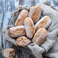 Die Dinkelchen liegen in einem mit einem grob gewebtem grauen Tuch ausgelegtem Drahtkorb. Bei einem auseinandergebrochenen Brötchen wird die kleinporige, elastische Krume sichtbar.