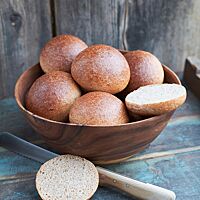 Die glänzenden, goldbraun ausgebackenen Burgerbuns liegen in einer Holzschale. Ein aufgeschnittener Burgerbun lässt die wattige, kleinporige Krume erkennen, die von einer feinen Kruste umgeben ist.