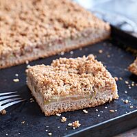 Der angeschnittene Blechkuchen liegt auf einem schwarzen Backblech. Im Anschnitt ist eine Schichtung des feinporigen Bodens, der saftigen Apfelschicht und der goldbraunen Streusel deutlich erkennbar.
