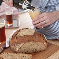 Neben dem angeschnittenen Weizenmischbrot mit kräftiger Kruste und lockerer Krume wird eine Flasche Bier in ein Glas eingeschenkt.
