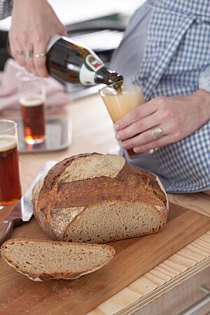 Neben dem angeschnittenen Weizenmischbrot mit kräftiger Kruste und lockerer Krume wird eine Flasche Bier in ein Glas eingeschenkt.
